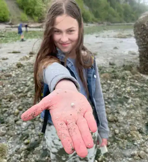 West Sound Academy Belong Deeply Smiling student reaching a hand toward the camera on a rocky beach shoreline