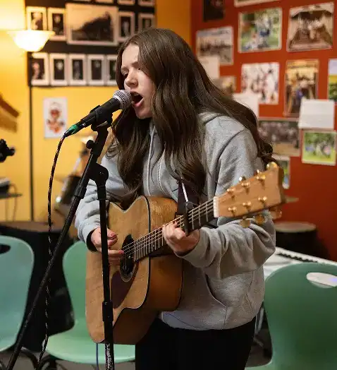 West Sound Academy Create with Purpose Student singing into a microphone while playing acoustic guitar in a room decorated with photos and artwork