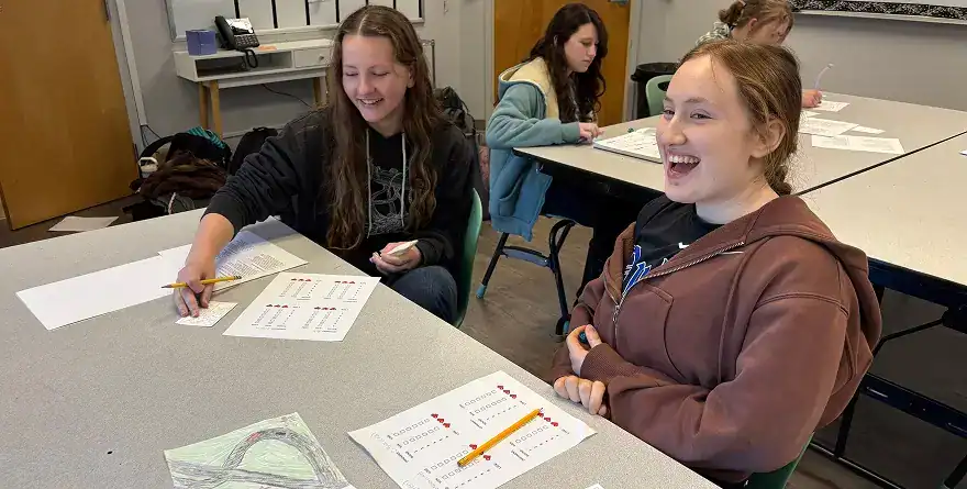 West Sound Academy Diploma Programme Two students laughing together at a desk with worksheets during class